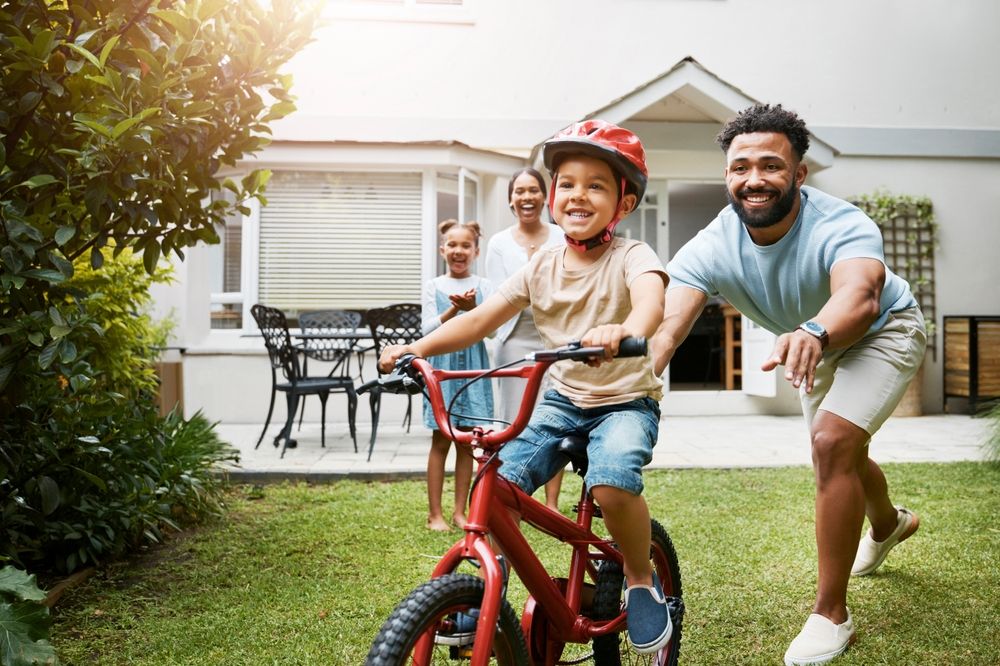 Learning, bicycle and proud dad teaching his young son to ride while wearing a helmet for safety in their family home garden.